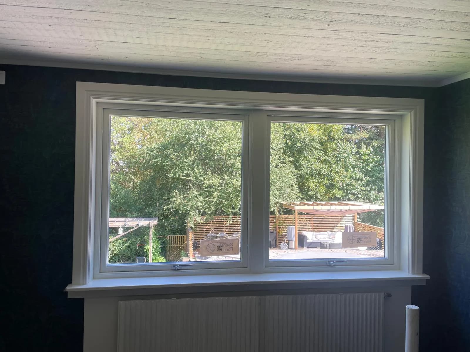 Två vita vridfönster sida vid sida i gemensam karm med profilerat foder mot mörk vägg, grönska utanför.