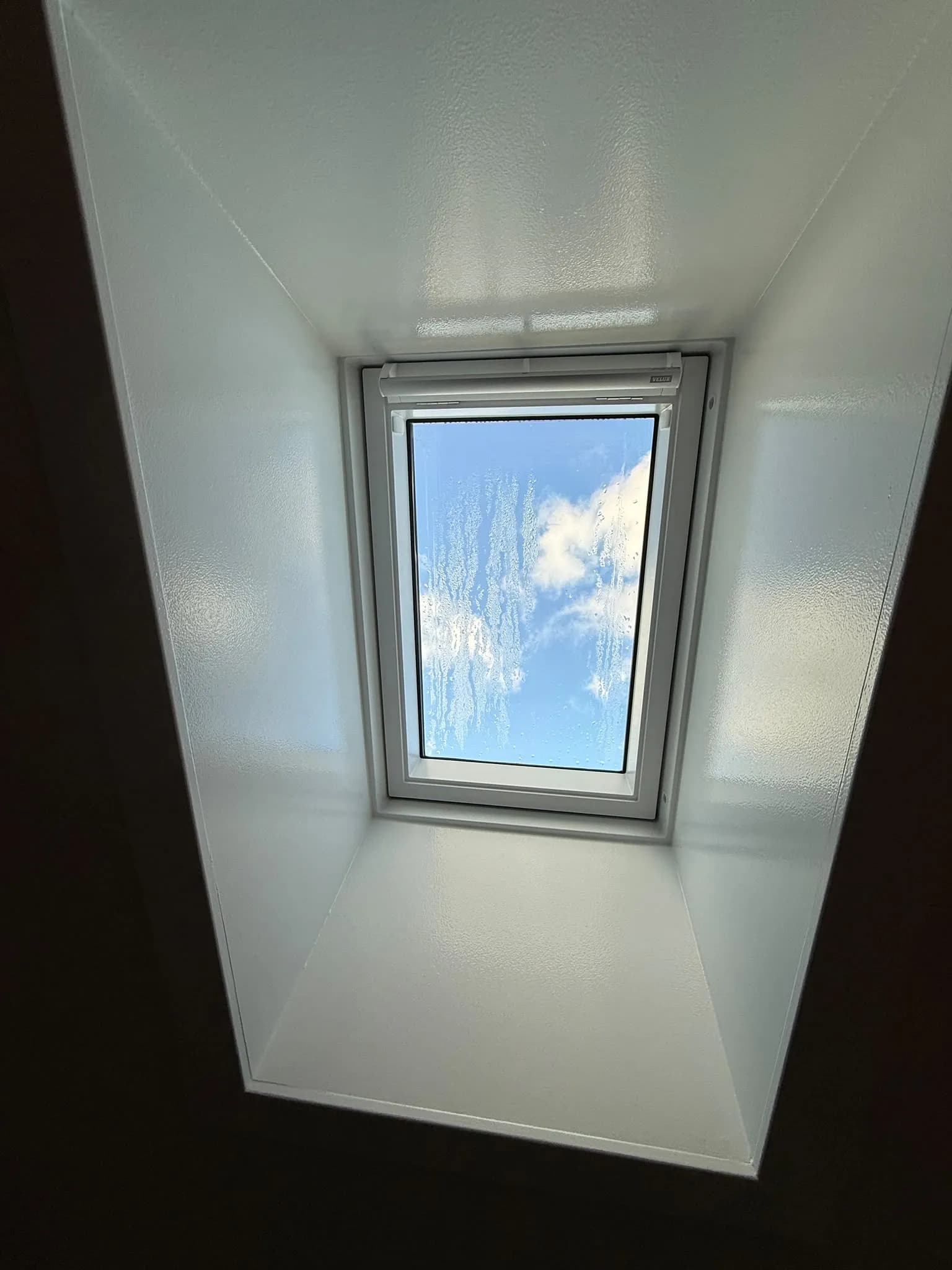 Takfönster sett rakt underifrån genom djup vit smyg, med rullgardinskassett och blå himmel med moln.