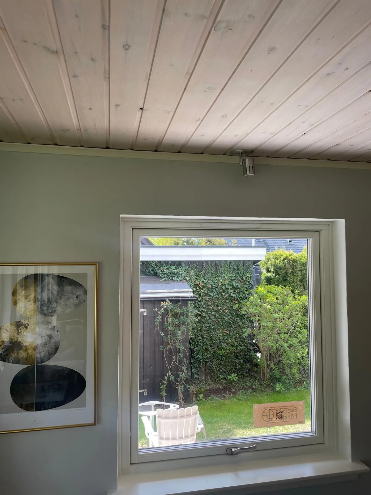 Vitt vridfönster inifrån under panelat tak, med djup smyg och fönsterbänk, utsikt mot gräsmatta och förråd.