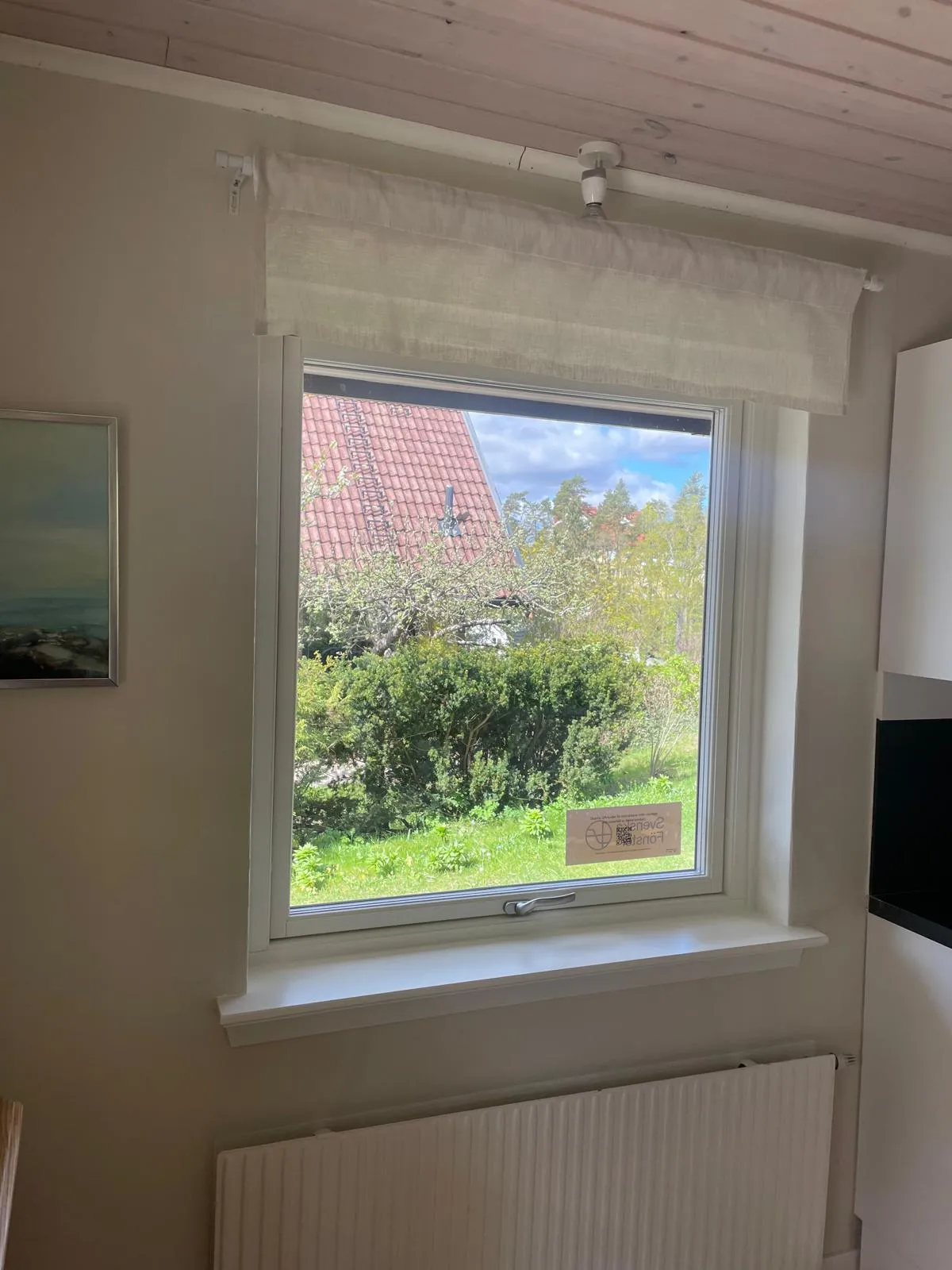 Vitt vridfönster inifrån i kök med vit fönsterbänk och släta smygar, hissgardin ovanför och radiator under.