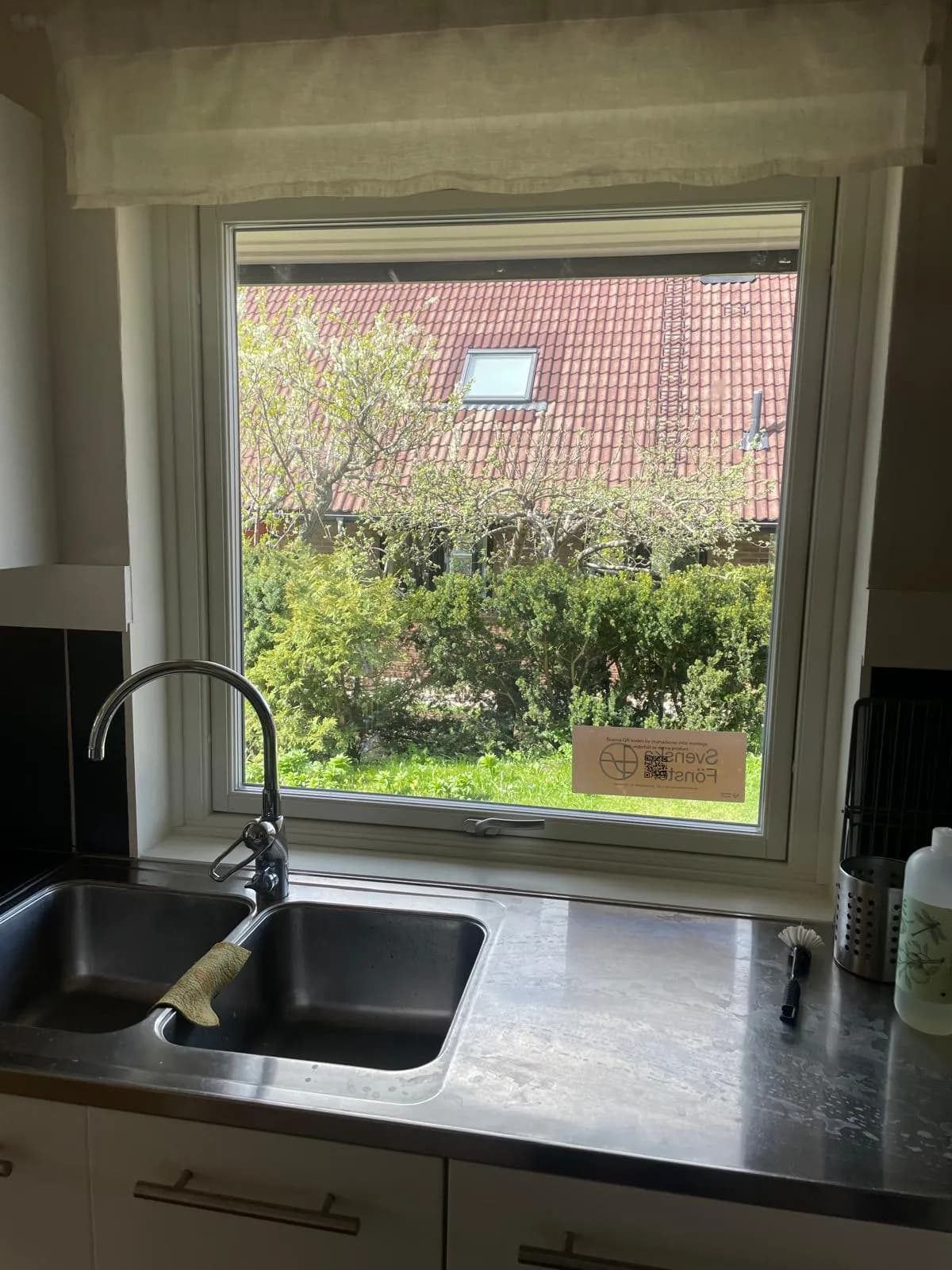 Vitt fönster ovanför diskbänk i rostfritt stål, med blandare i förgrunden och trädgård utanför.