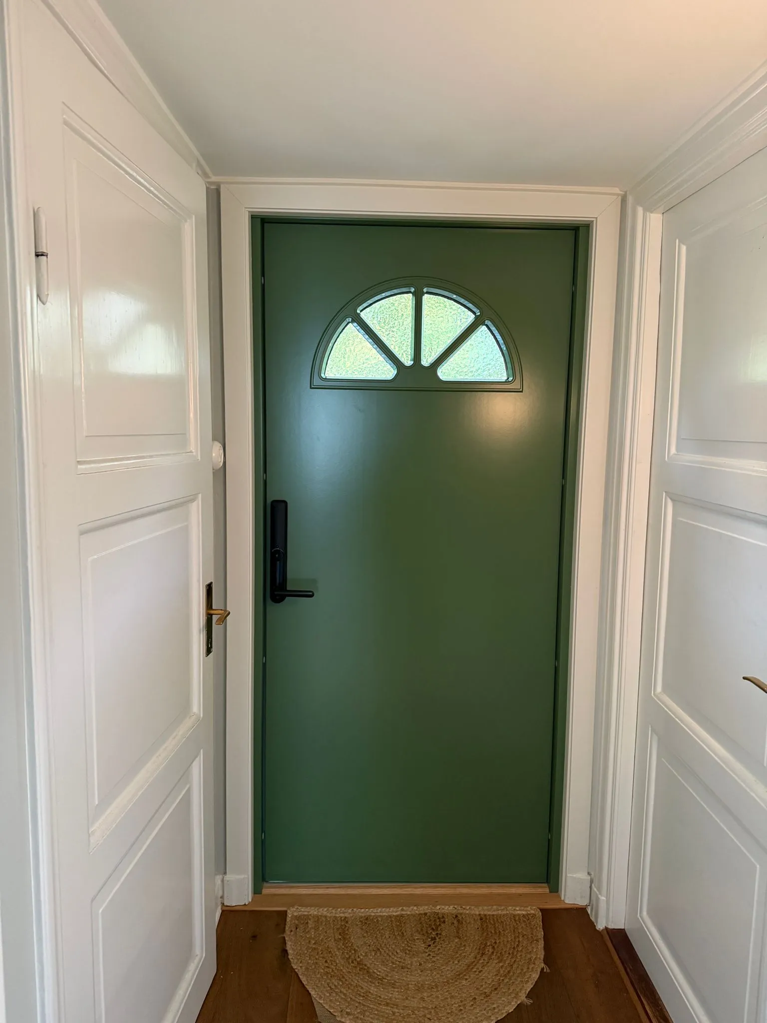 Insidan av en grön ytterdörr med halvmåneformat glas och svart Yale Doorman, mellan vita spegeldörrar i en ljus hall.