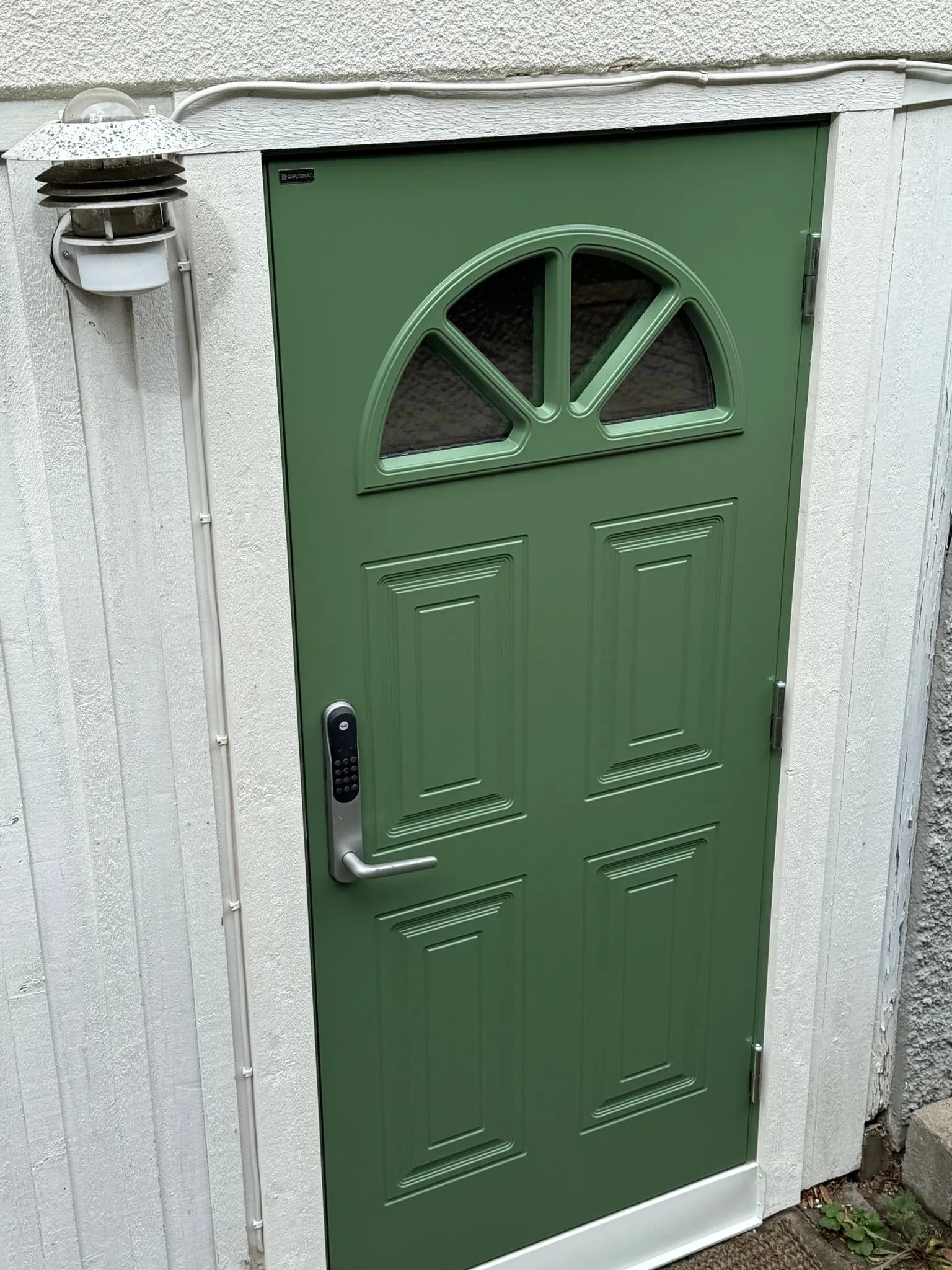 Närbild utifrån på grön fyllningsdörr med halvmåneformat glasparti och Yale Doorman med kodlås, bredvid en vägglampa på vit fasad.